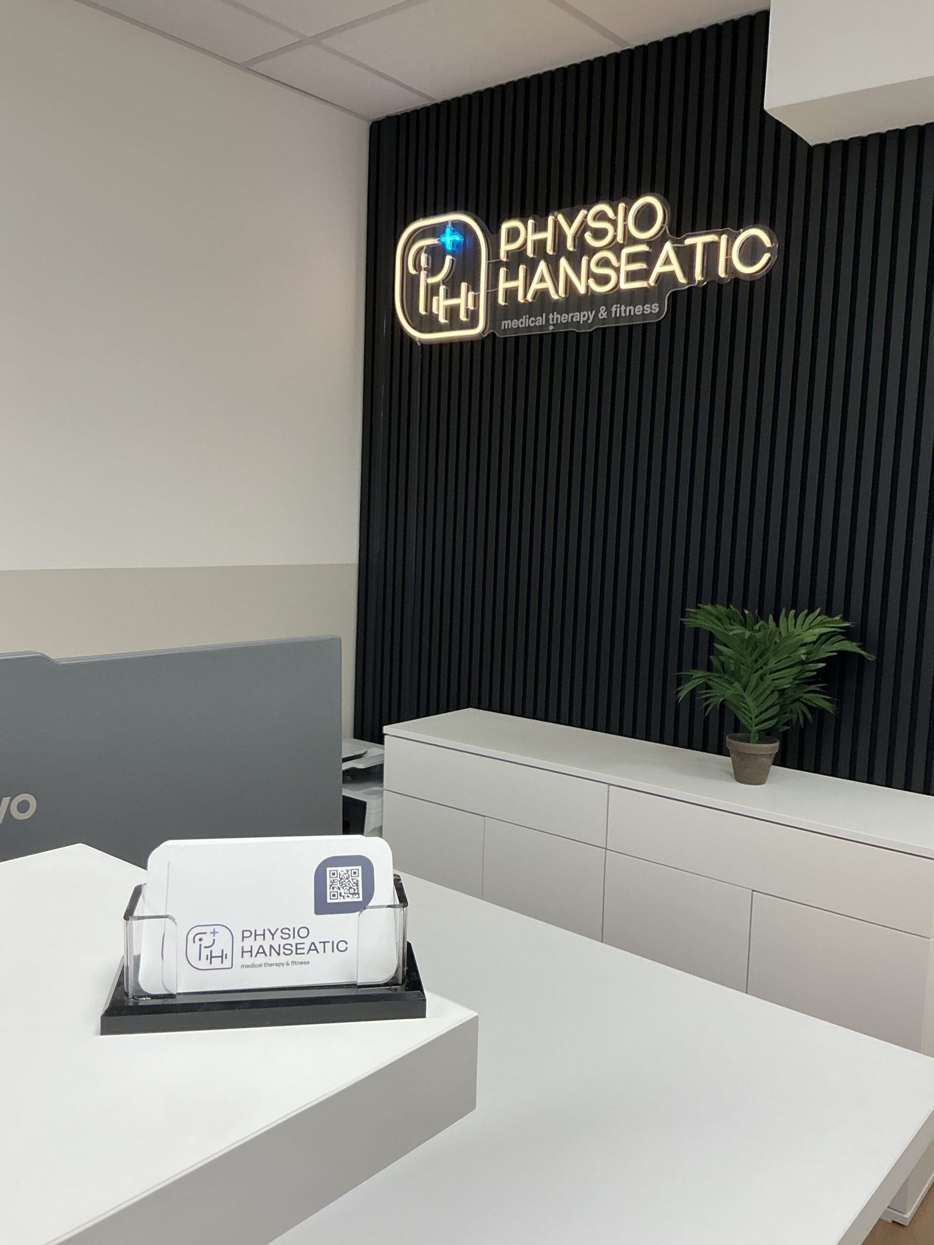 Empfangstresen mit Visitenkartenhalter und beleuchtetem Logo "PHYSIO HANSEATIC medical therapy & fitness" an schwarzer Wand mit vertikalen Lamellen und einer Pflanze auf einem weißen Sideboard