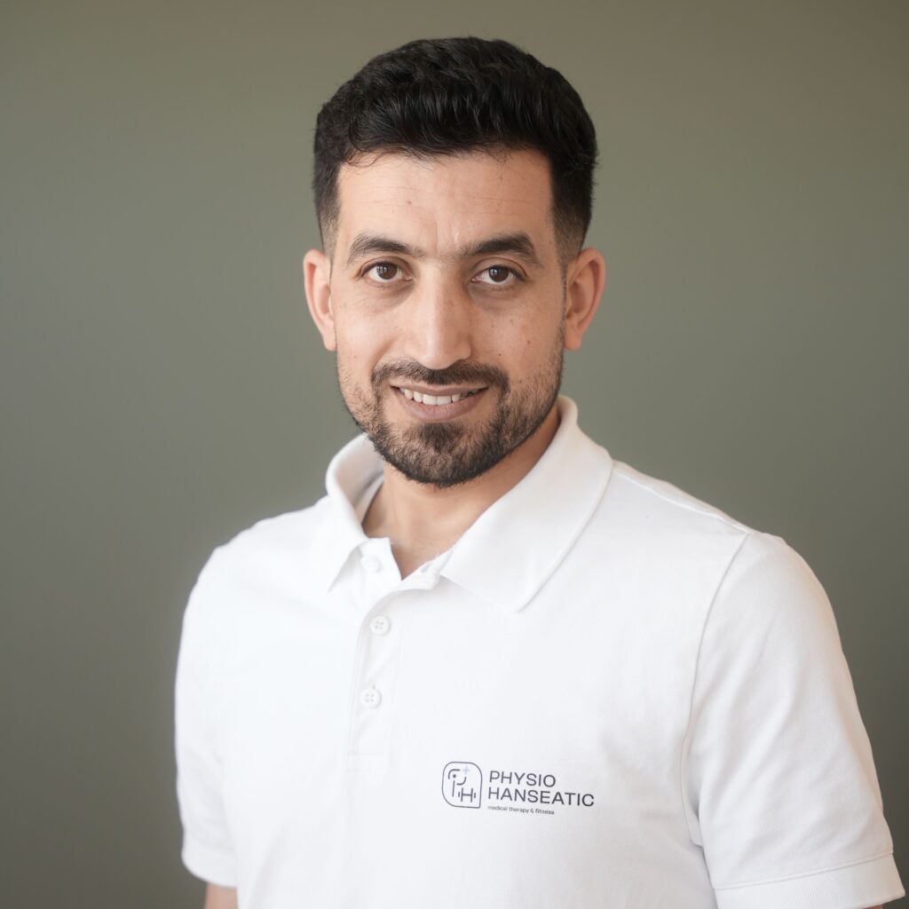 Faisal Hamdo mit kurzem lockigem Haar und Bart in weißem Poloshirt mit Logo steht vor grauer Wand und lächelt.