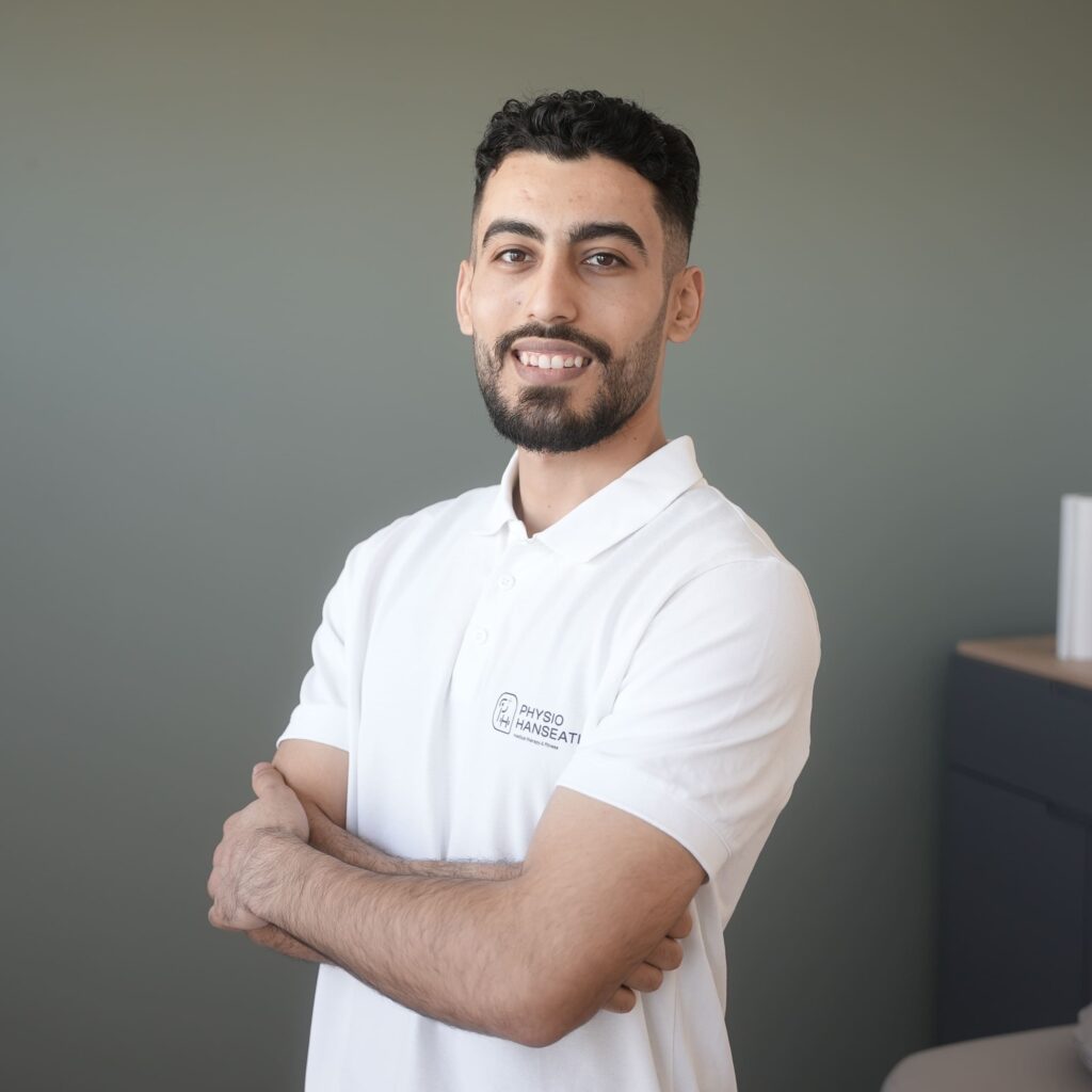 Sleiman Hamdo mit kurzem lockigem Haar und Bart in weißem Poloshirt mit Logo steht vor grauer Wand und lächelt mit verschränkten Armen