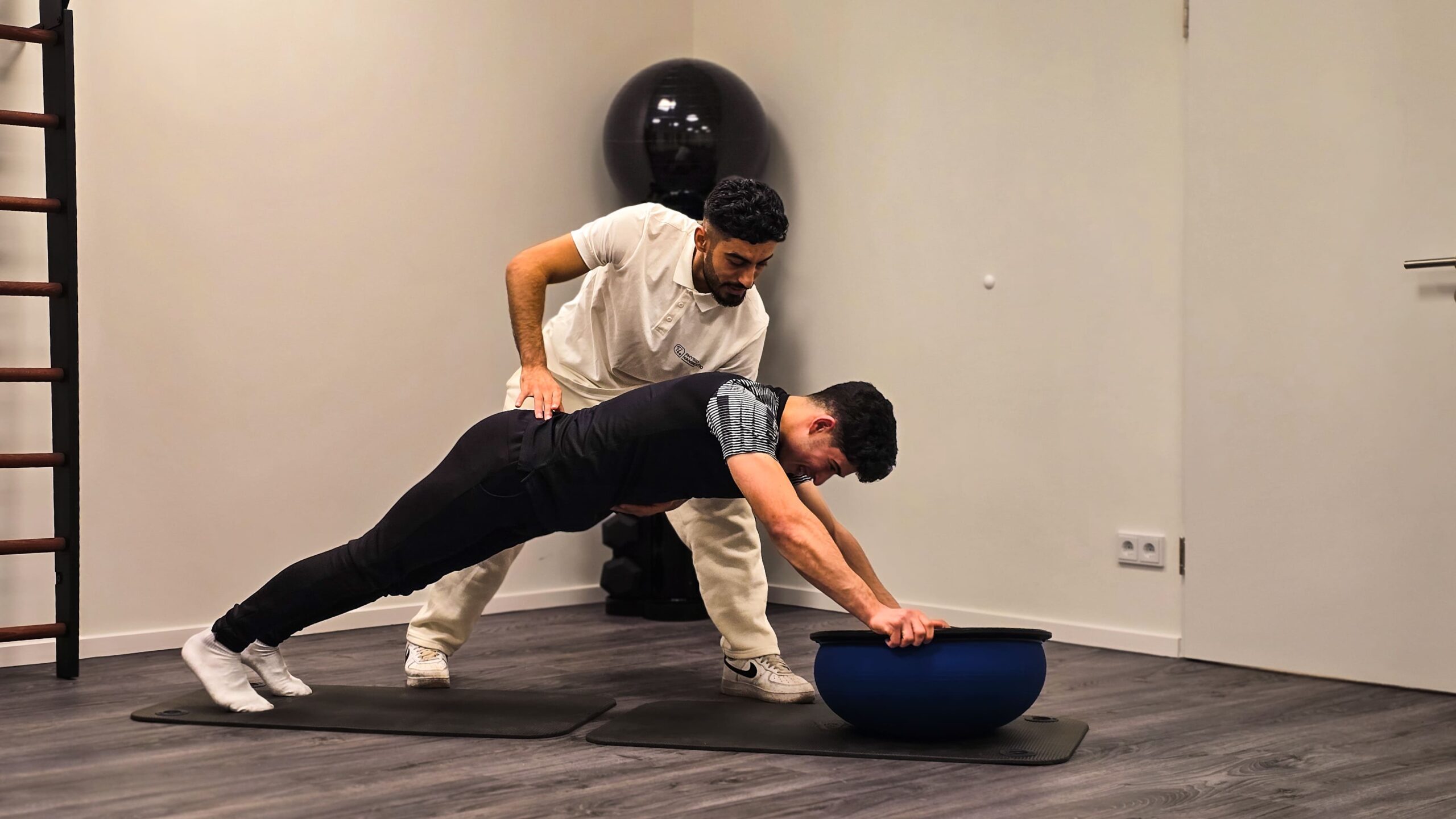 Zwei Männer in einem Behandlungsraum, einer stützt sich in Liegestützposition auf einem halbrunden Balancegerät ab, der andere hält ihn an der Hüfte zur Stabilisierung